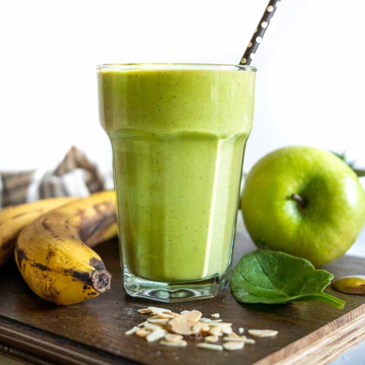 A glass of green smoothie surrounded by bananas, apples, and almond flakes