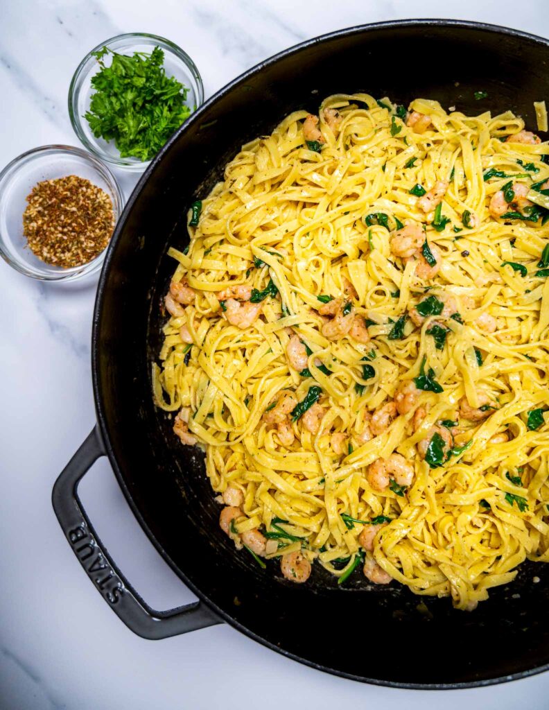 Close up of a pot of pasta with shrimp and spinach with small bowls of zaatar and parsley