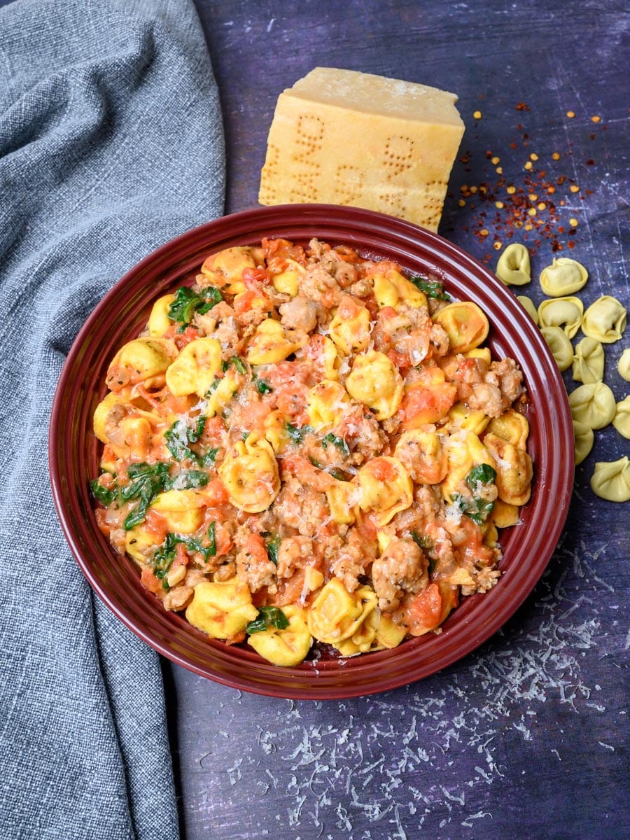 A red bowl of pasta with a block of Parmesan on the side.