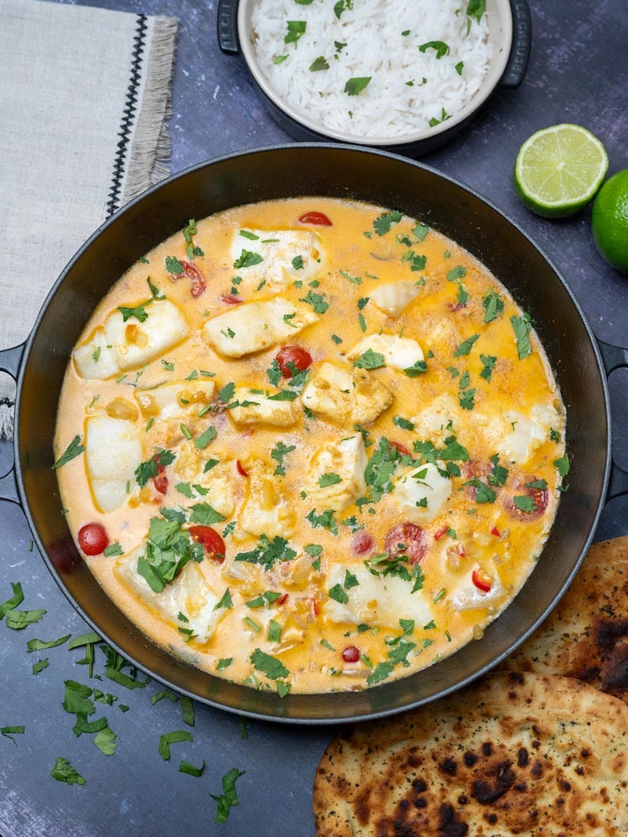Overhead view of creamy white fish in golden curry sauce served with lime wedges and fresh coriander