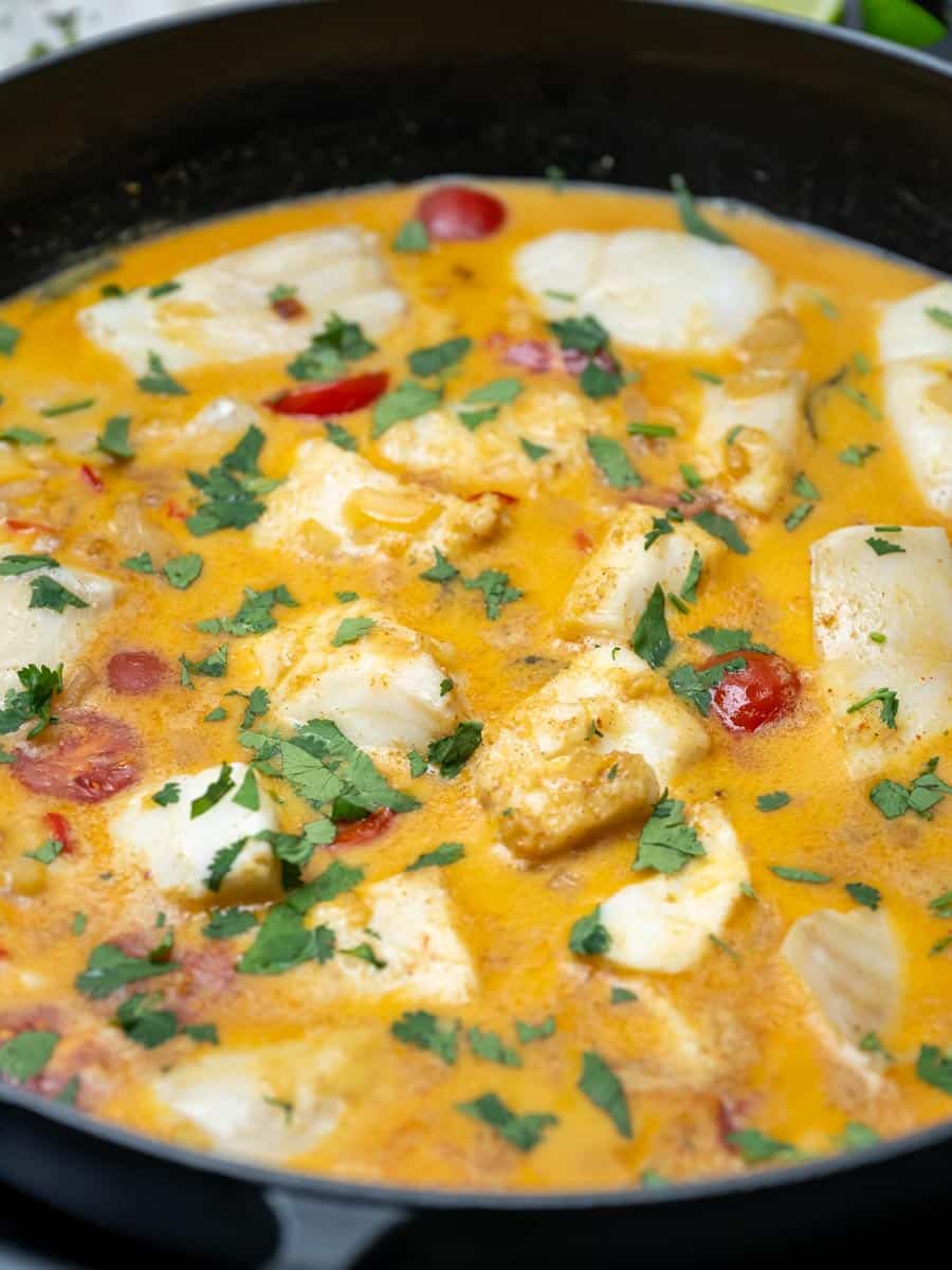 Close-up of creamy fish curry with chunks of white fish, cherry tomatoes, and fresh coriander in a rich golden sauce