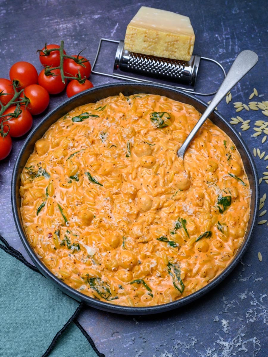 A bowl of creamy tomato orzo with a chunk of Parmesan and cherry tomatoes on the side