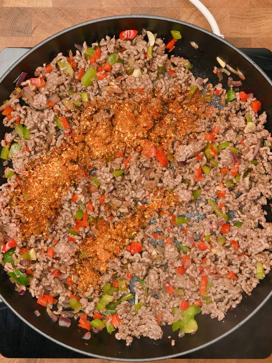 Adding ground beef and taco seasoning to a black skillet with veggies