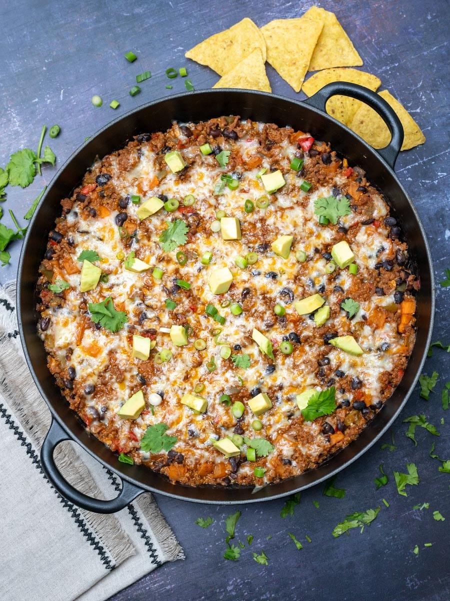 A photo of a black skillet with Tex-Mex ground beef casserole topped with avocados and with tortilla chips on the side