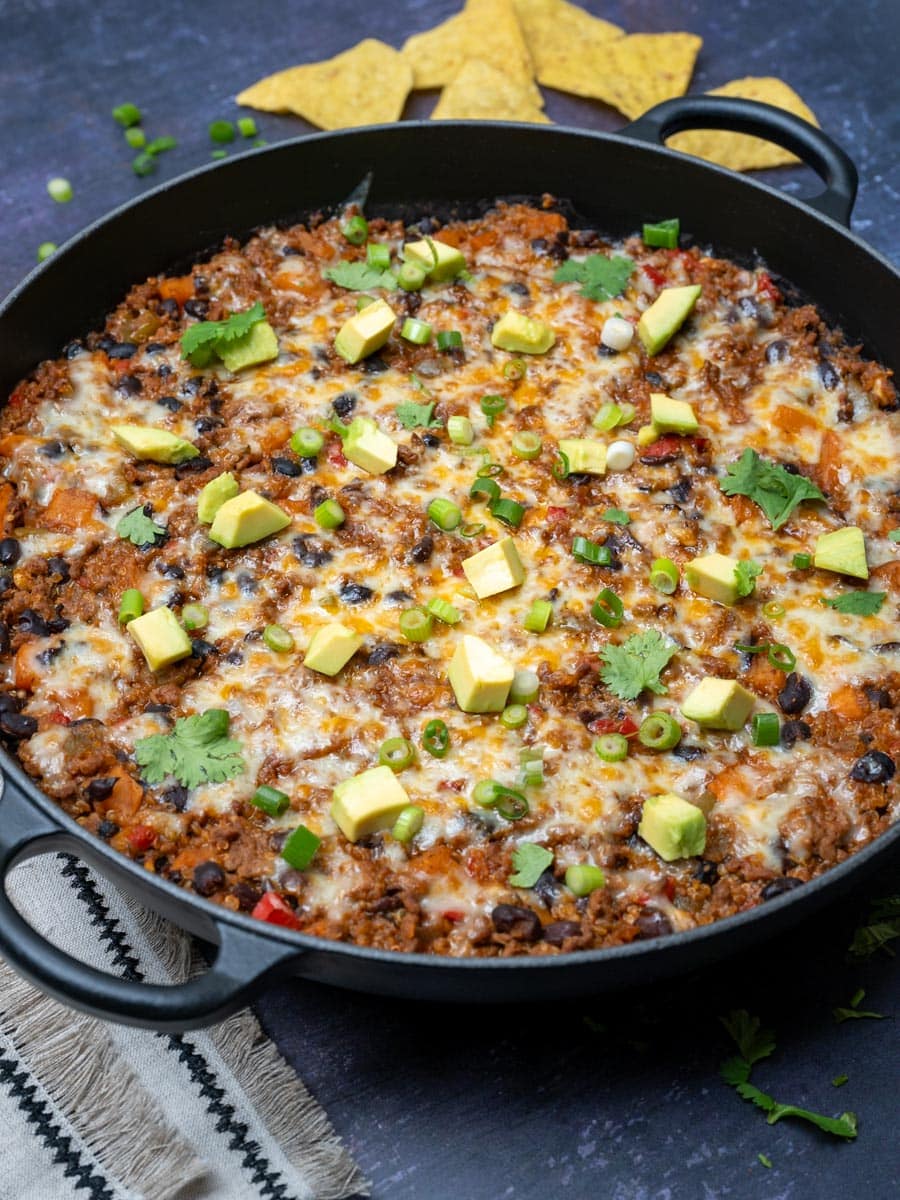 A close up photo of a Mexican-style skillet with ground beef and beans, topped with avocado, scallions and cilantro