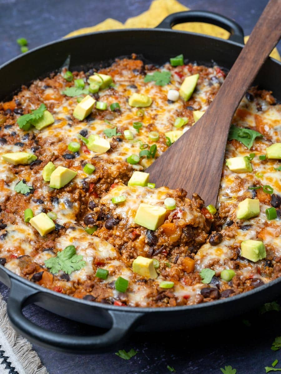 A close up photo of a Mexican ground beef skillet with freshly chopped cilantro, scallions and avocado with a wooden spoon in it